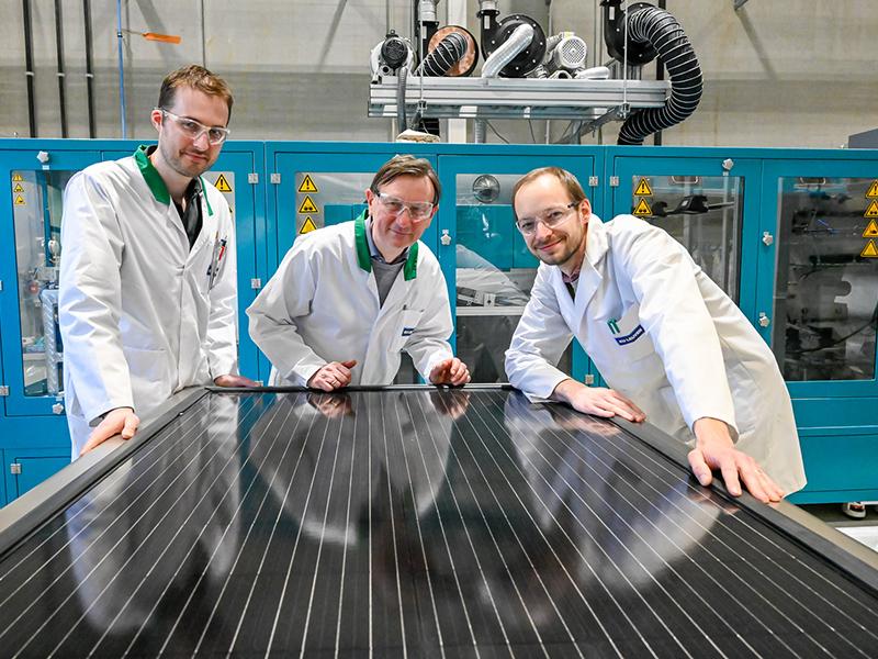 Three male inventors in white cloaks, in front of big solar panel in construction hall