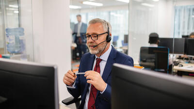 Bearded and bespectacled middle-aged man wearing a suit and tie speaking into a headset while sitting at a desk and looking at a computer screen, holding a pen in both hands