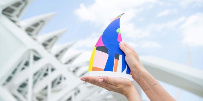 A hand holding a colorful European Inventor Award trophy with the Ciutat de les Arts of Valencia on the background